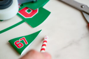 Hand applying a loop of tape to the top of a red-and-white straw to attach the small green &ldquo;67&rdquo; pennant for the Elf on the Shelf setup.