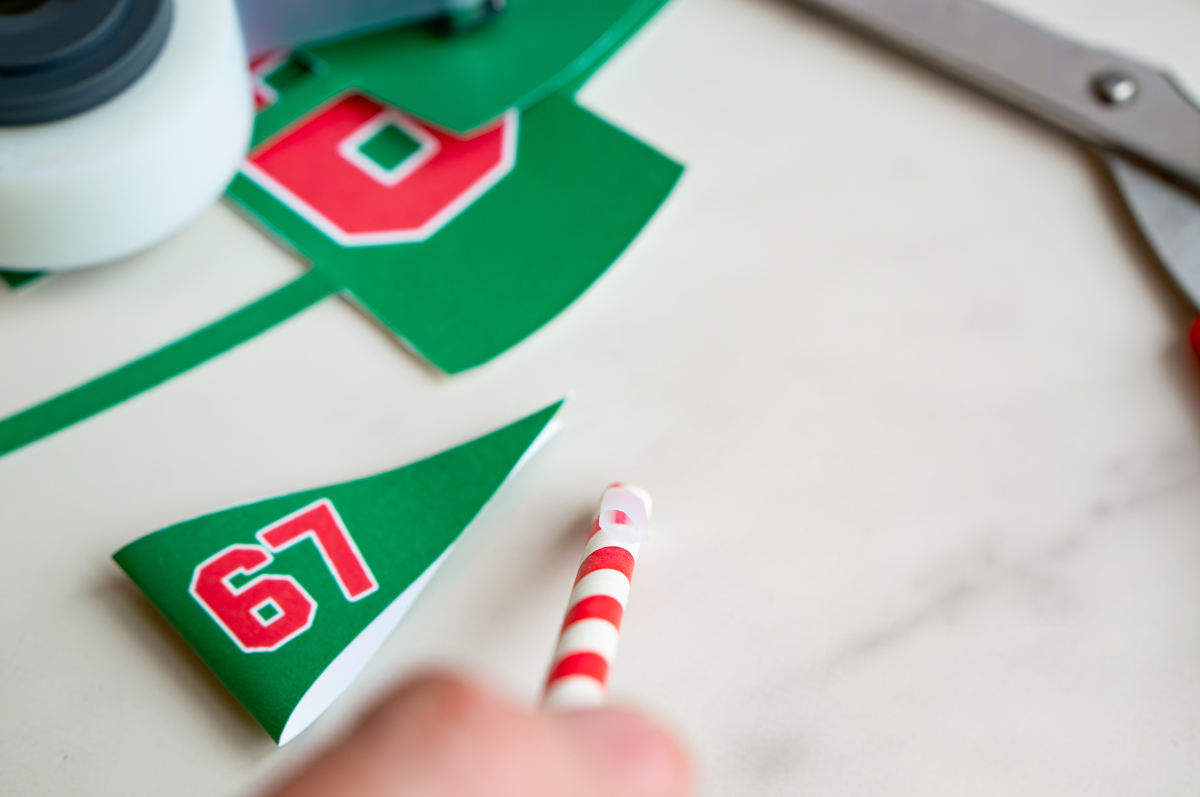 Hand applying a loop of tape to the top of a red-and-white straw to attach the small green &ldquo;67&rdquo; pennant for the Elf on the Shelf setup.