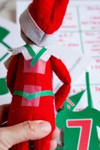Back view of an Elf on the Shelf doll showing how the green jersey straps are secured with clear tape for easy attachment.