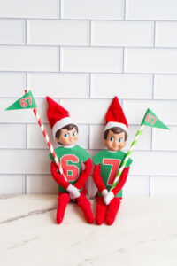 Two Elf on the Shelf dolls wearing green paper jerseys numbered 6 and 7, each holding a festive striped straw with a matching &ldquo;67&rdquo; pennant.