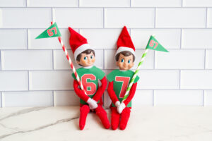 Two Elf on the Shelf dolls sitting together wearing jerseys numbered 6 and 7 and holding coordinating pennant flags on striped straws.