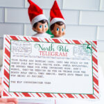 A hand holds up a &ldquo;Sorry I&rsquo;m Late&rdquo; North Pole telegram with a candy-cane striped border, with two Elf on the Shelf dolls blurred in the background.