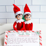 Two Elf on the Shelf dolls sit behind a printed &ldquo;Sorry I&rsquo;m Late&rdquo; North Pole telegram with a red-and-white candy cane border, displayed on a marble countertop.
