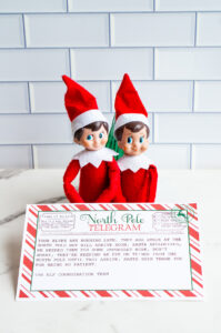 Two Elf on the Shelf dolls sit behind a printed &ldquo;Sorry I&rsquo;m Late&rdquo; North Pole telegram with a red-and-white candy cane border, displayed on a marble countertop.