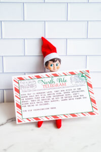 An Elf on the Shelf doll peeks over the top of a &ldquo;Sorry I&rsquo;m Late&rdquo; North Pole telegram printable, propped against a white subway-tile wall.
