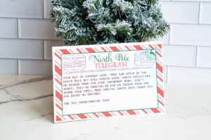 A &ldquo;Sorry I&rsquo;m Late&rdquo; North Pole telegram printable stands upright in front of a small frosted Christmas tree on a marble surface.