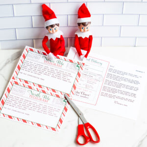 Two Elf on the Shelf dolls sit on a white countertop behind printed North Pole telegram letters and an sorry I'm late letter. A pair of red scissors rests beside the printables.