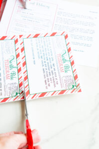 A close-up of a hand using red scissors to cut out a North Pole telegram printable, with an Elf on the Shelf sorry I'm late letter visible in the background.
