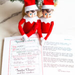 Two Elf on the Shelf dolls sit in front of a small frosted Christmas tree, resting their hands on a printed North Pole sorry I'm late letter addressed to Carl and Jack.
