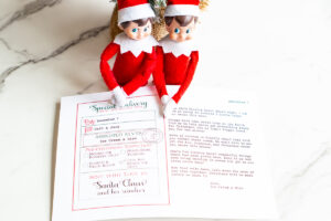 Two Elf on the Shelf dolls sit above a North Pole sorry I'm late letter laid out on a marble surface, showing assigned elves and a typed sorry I'm late letter.