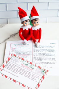 Two Elf on the Shelf dolls sit behind both a North Pole &ldquo;Sorry I&rsquo;m Late&rdquo; letter and a late-arrival telegram, arranged together on a white countertop.