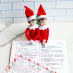 Two Elf on the Shelf dolls sit behind a customizable &ldquo;Sorry I&rsquo;m Late&rdquo; North Pole letter and telegram printable laid out on a marble surface.