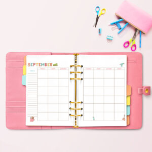 Open pink planner displaying the September craft-themed undated calendar, surrounded by colorful scissors, binder clips, and a pink pencil pouch with craft supplies.