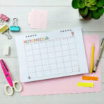 November craft-themed undated calendar in a spiral-bound format, laid on a desk with binder clips, pens, pencils, and a small succulent plant.