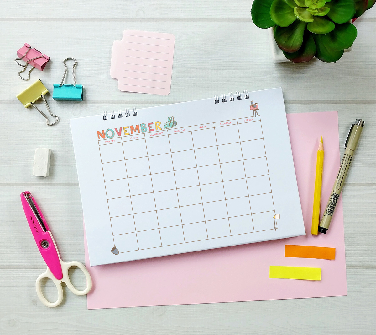 November craft-themed undated calendar in a spiral-bound format, laid on a desk with binder clips, pens, pencils, and a small succulent plant.