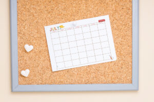 July craft-themed undated calendar pinned to a corkboard with two heart-shaped magnets placed beside it.