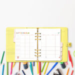 Open yellow planner showing the September craft-themed undated calendar, placed above scattered colored pencils, markers, and craft tools.