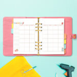 Open pink planner displaying the March craft-themed undated calendar, with fabric scissors, a glue gun, yellow fabric, and craft tools arranged around it.