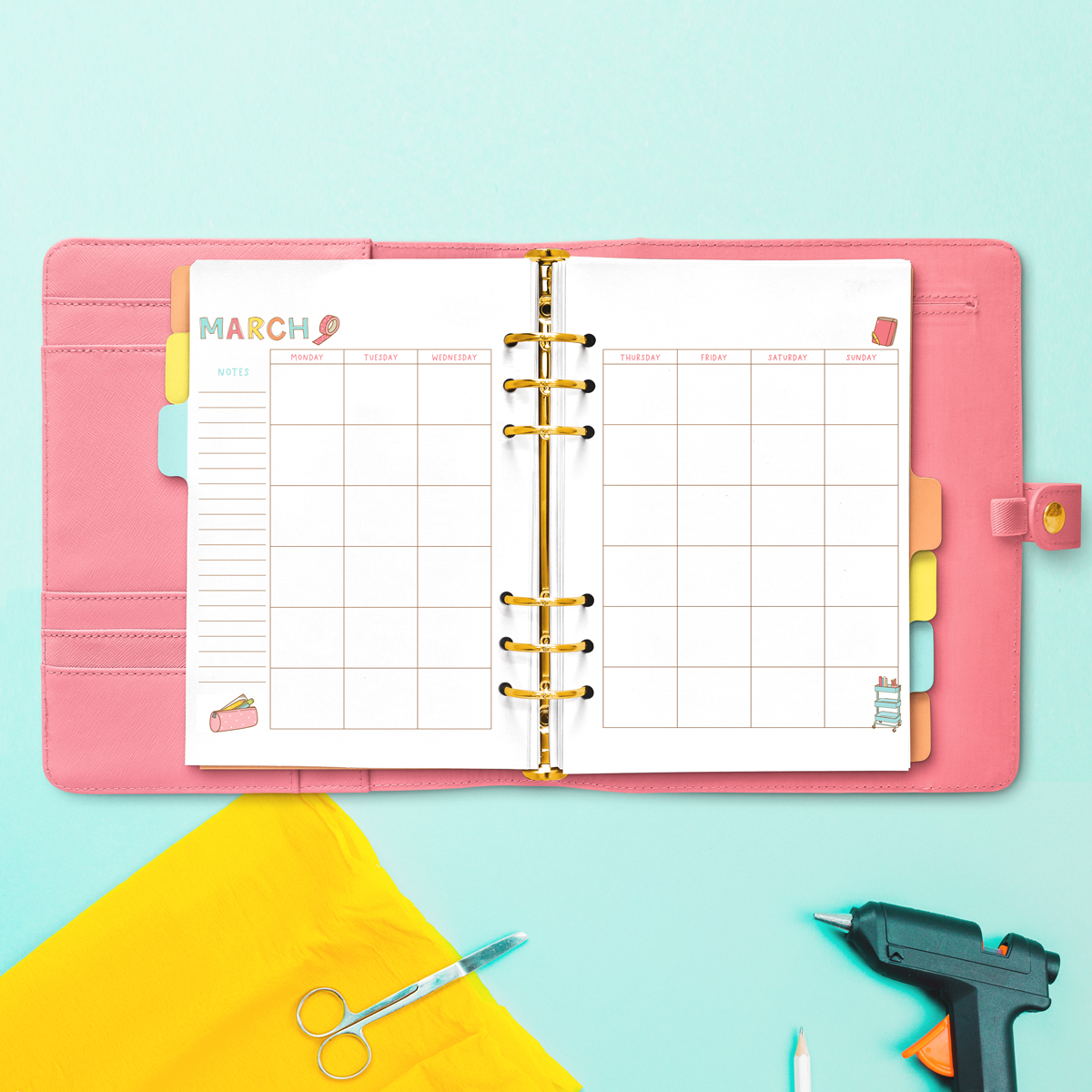 Open pink planner displaying the March craft-themed undated calendar, with fabric scissors, a glue gun, yellow fabric, and craft tools arranged around it.