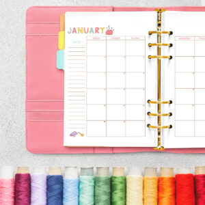 Open pink planner featuring the January craft-themed undated calendar, with a row of colorful embroidery thread spools lined up at the bottom.