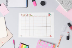 August craft-themed undated calendar page surrounded by notebooks, sticky notes, pens, highlighters, and makeup items on a gray background.