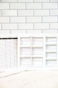 A photo of three printable contact list pages standing upright against a white subway-tile background. The left page features a black-topped column layout with rows for names, two phone numbers, and email. The middle and right pages show boxed contact-card layouts in muted colors with spaces for name, phone numbers, email, address, notes, and socials.