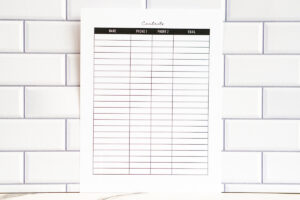 A full-page view of the lined contact list layout with a black header row labeled Name, Phone 1, Phone 2, and Email. The sheet is displayed upright in front of a white subway-tile wall.