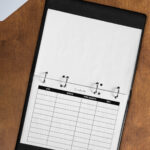 A lined contact list page inserted in a black leather-style binder on a wooden desk, shown alongside a laptop and a cup of tea.