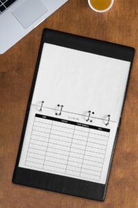 A lined contact list page inserted in a black leather-style binder on a wooden desk, shown alongside a laptop and a cup of tea.