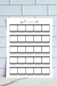 A printed movie tracker page displayed against a light blue tile wall. The sheet features horizontal film-strip frames with blank boxes for movie titles and star-rating rows under each frame.