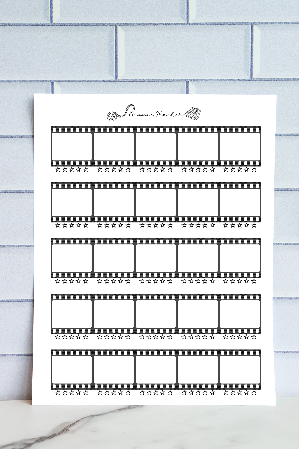 A printed movie tracker page displayed against a light blue tile wall. The sheet features horizontal film-strip frames with blank boxes for movie titles and star-rating rows under each frame.
