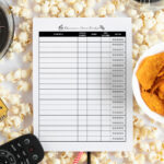 Printed TV show tracker sheet placed on a bed of popcorn, surrounded by a TV remote, sunglasses, a film reel, and a bowl of chips.