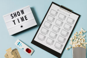 Flatlay of a TV show tracker printable on a clipboard, styled with a &ldquo;SHOW TIME&rdquo; lightbox, vintage cinema tickets, 3D glasses, and a striped container of popcorn on a light blue background.