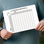 Person in a teal long-sleeve shirt holding a printed black-and-white football squares chart in front of their torso.