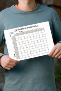 Person in a teal long-sleeve shirt holding a printed black-and-white football squares chart in front of their torso.