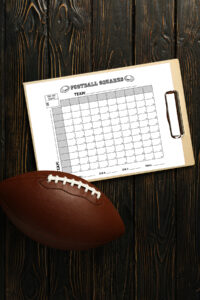Football squares printable displayed on a clipboard resting on dark wooden boards with a brown football placed in the foreground.