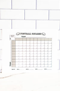 Football squares printable propped against a white subway-tile backsplash with the grid and team labels clearly visible.