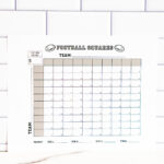 Close-up of the football squares printable leaning against a subway-tile wall, highlighting the team lines and numbered grid.