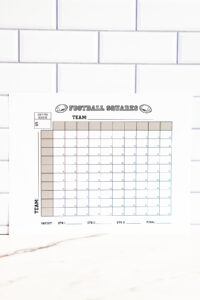 Close-up of the football squares printable leaning against a subway-tile wall, highlighting the team lines and numbered grid.