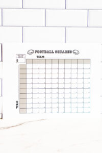 Football squares printable positioned on a marble countertop with a subway-tile wall behind it, showing the empty squares and team labels.