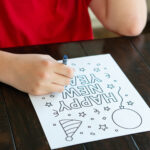 A child wearing a red shirt colors a &ldquo;Happy New Year&rdquo; coloring page at a dark wooden table. The page features festive outlines including stars, confetti, a balloon, and a party hat.