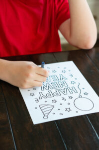 A child wearing a red shirt colors a &ldquo;Happy New Year&rdquo; coloring page at a dark wooden table. The page features festive outlines including stars, confetti, a balloon, and a party hat.