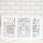 Three &ldquo;Happy New Year&rdquo; coloring pages displayed side by side against a white subway-tile wall, featuring designs with balloons, an axolotl in a party hat, and a bear surrounded by festive decorations.