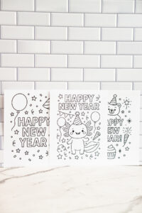 Three &ldquo;Happy New Year&rdquo; coloring pages displayed side by side against a white subway-tile wall, featuring designs with balloons, an axolotl in a party hat, and a bear surrounded by festive decorations.