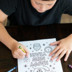 A child in a black shirt colors a &ldquo;Happy New Year&rdquo; bear-themed coloring page at a dark wooden table. Crayons are scattered around as the child fills in fireworks and festive elements.