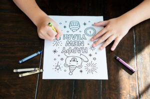 A child colors a &ldquo;Happy New Year&rdquo; bear coloring page at a wooden table, using a yellow crayon. Crayons lie scattered nearby as sunlight falls across the workspace.