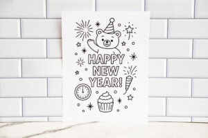 A printed &ldquo;Happy New Year&rdquo; coloring page taped against a white subway-tile backdrop, featuring a waving bear in a party hat surrounded by fireworks, stars, a cupcake, and a clock.
