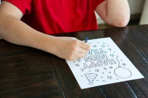 A child in a red shirt colors a &ldquo;Happy New Year&rdquo; coloring page upside down on a dark wooden table, with festive outlines of stars, confetti, a balloon, and a party hat.