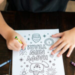 A child colors a &ldquo;Happy New Year&rdquo; coloring page featuring a bear, cupcake, fireworks, and a clock. Crayons are scattered nearby on a dark wooden table.