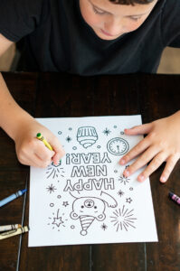 A child colors a &ldquo;Happy New Year&rdquo; coloring page featuring a bear, cupcake, fireworks, and a clock. Crayons are scattered nearby on a dark wooden table.